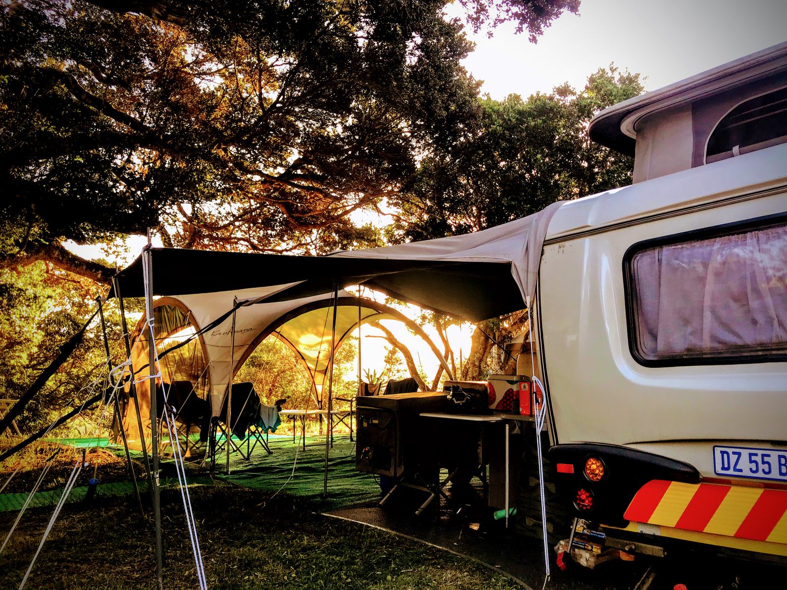 A caravan and awning pitched under coastal trees at golden hour
